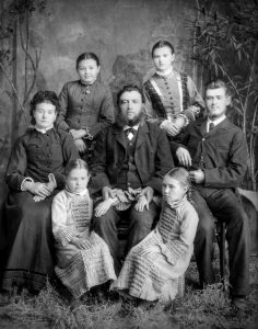 A photo of a family featuring an older man with a chinstrap beard in the centre. He is surrounded by his four daughters, one older son, and his wife.